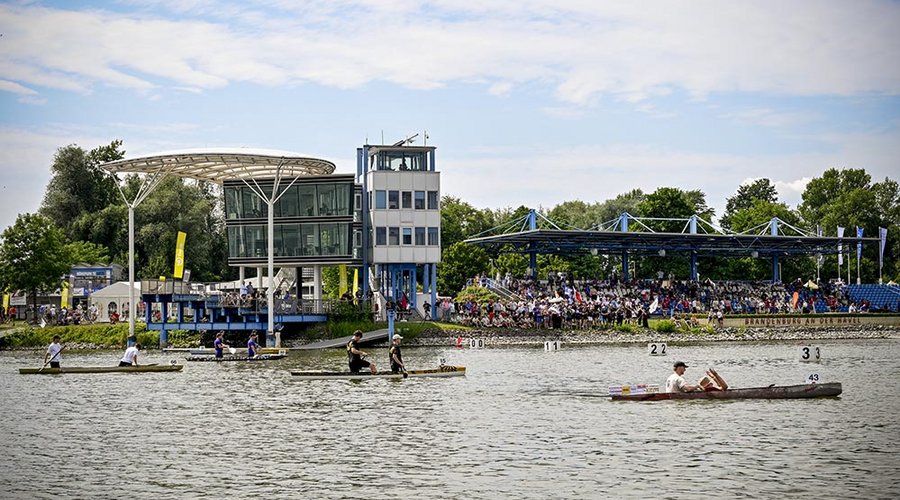 Ruderer auf einem See vor modernen Betonbauten und Tribünen mit Zuschauern bei e